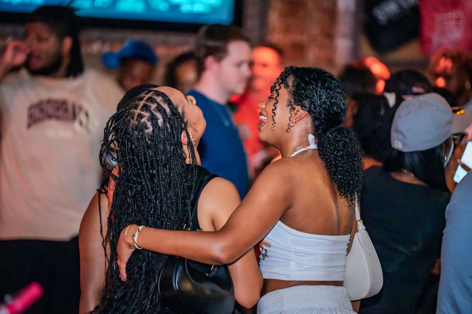 Two women hugging and laughing together on a crowded nightclub dance floor.