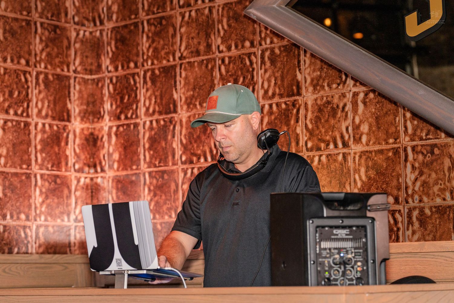 DJ in black shirt and cap mixing music at a booth with copper textured wall backdrop.