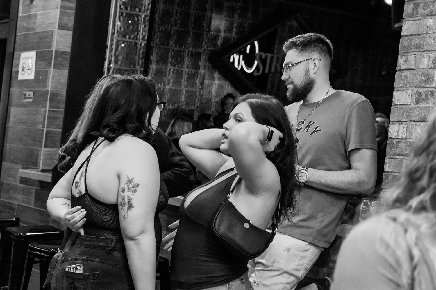 Black and white photo of two women and a bearded man socializing outside a brick building bar.