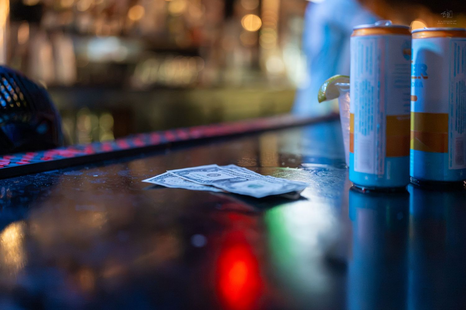 Cash and wallet on a dark bar counter beside two blue energy drink cans in a dimly lit nightclub.