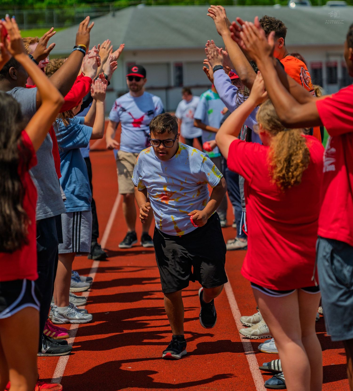 Athlete runs through cheering crowd tunnel on red track at Special Olympics event outdoors.