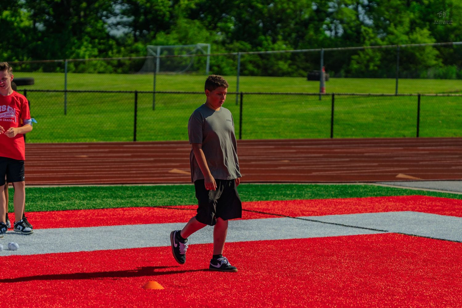 A young athlete walks across a red turf field during outdoor sports practice on a sunny day.
