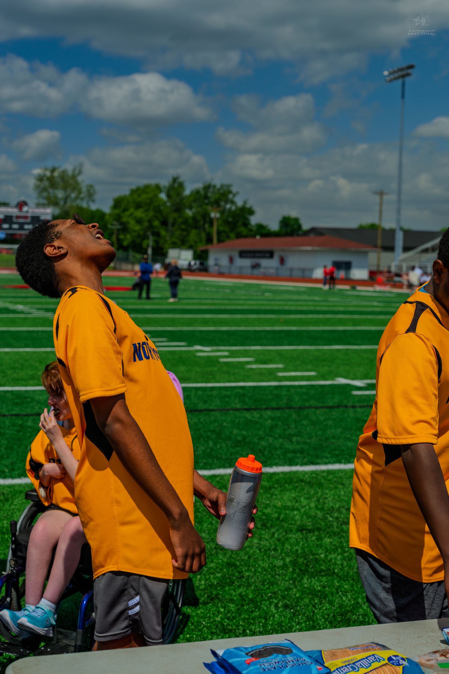 Young athletes in yellow jerseys stand near a table on a green turf field during an outdoor sports event under cloudy skies.