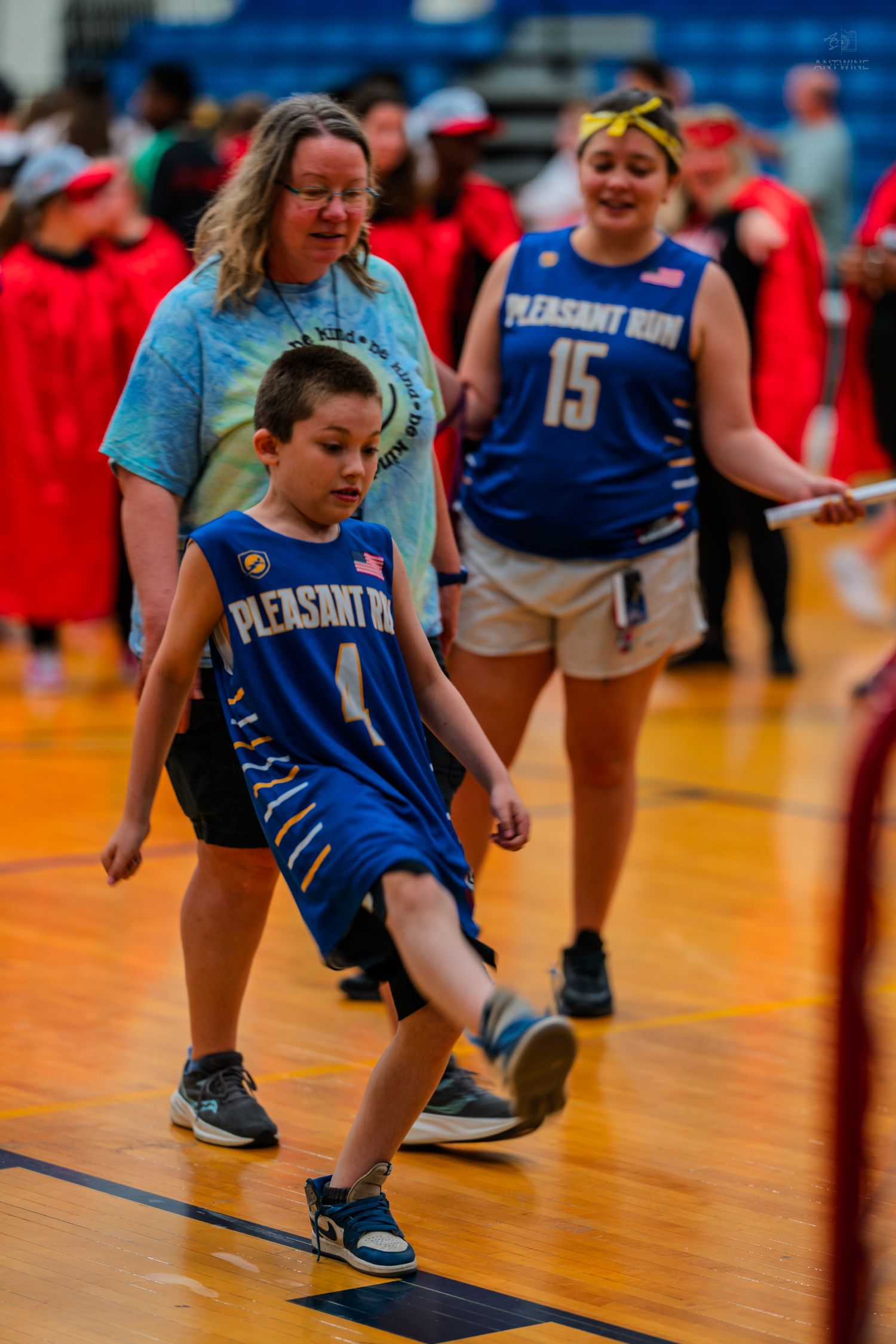 Young boy in Pleasant Run basketball jersey walks on gym floor with woman and teen girl in matching blue uniforms.