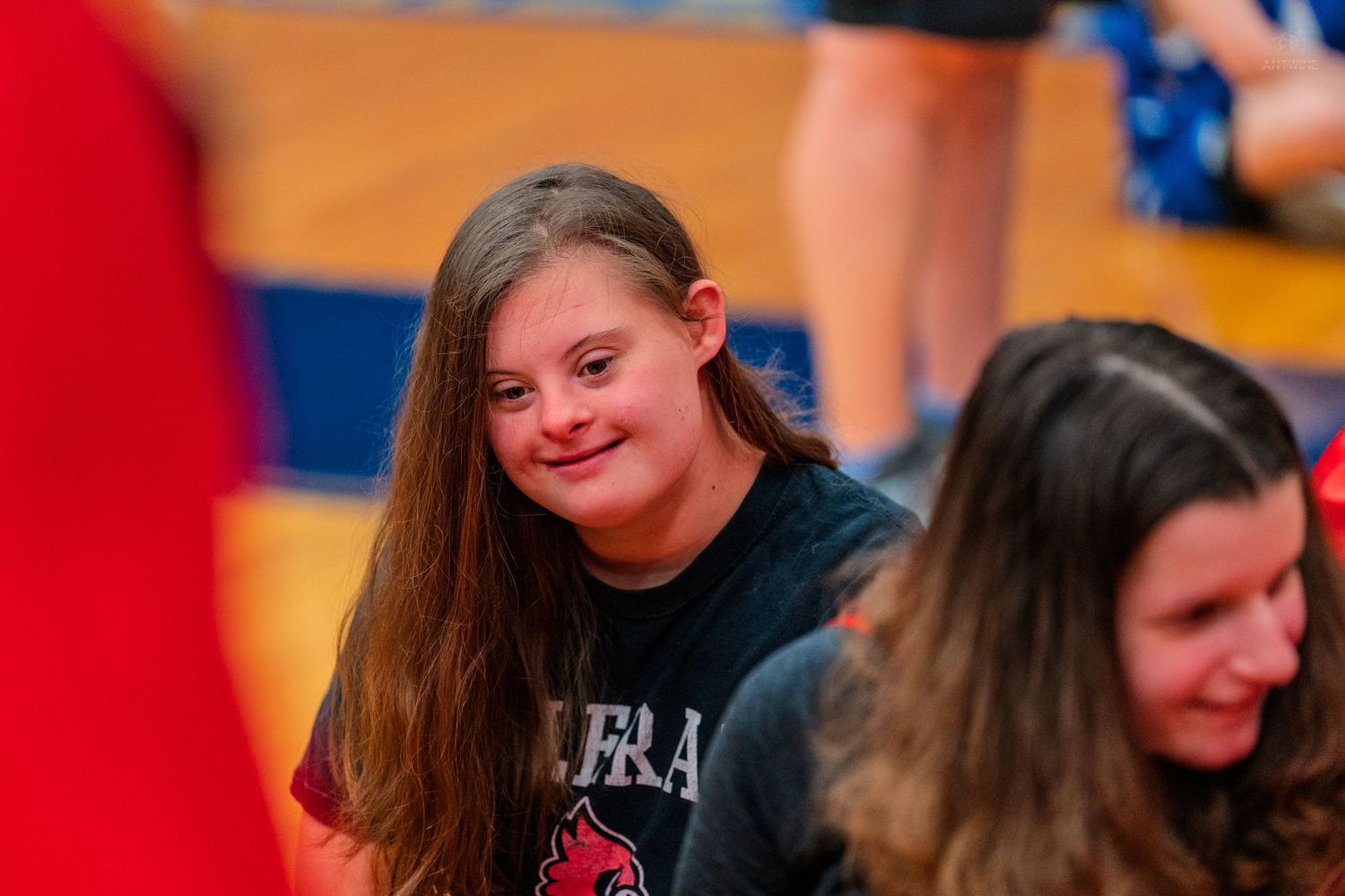 Smiling girl with Down syndrome at a school gym event wearing a red t-shirt.