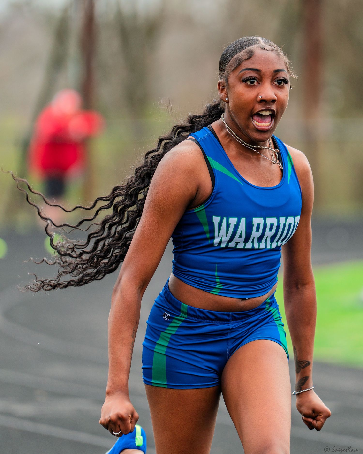 Female track athlete in Warriors uniform sprinting with hair flying during a relay race competition.