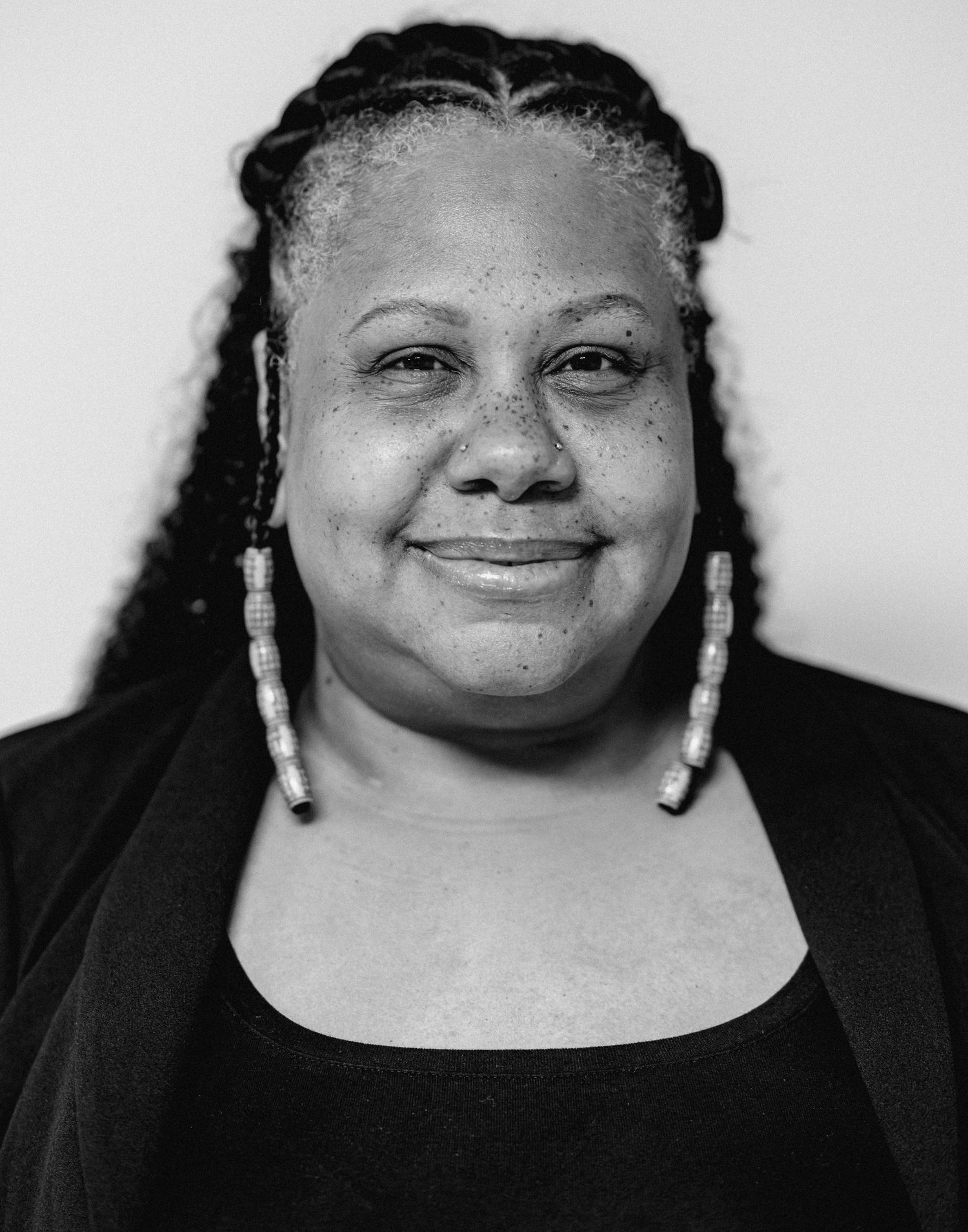 A Black woman with braided hair and beaded earrings smiles warmly in a black and white portrait photo.