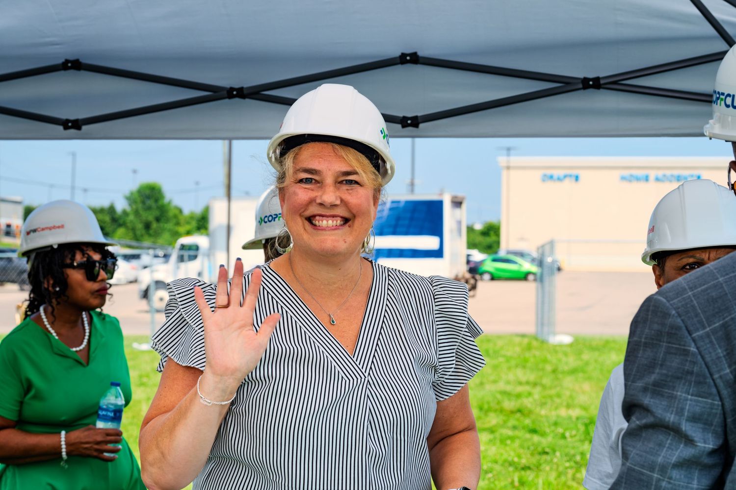 Smiling woman in white hard hat waving at an outdoor construction event under a tent.
