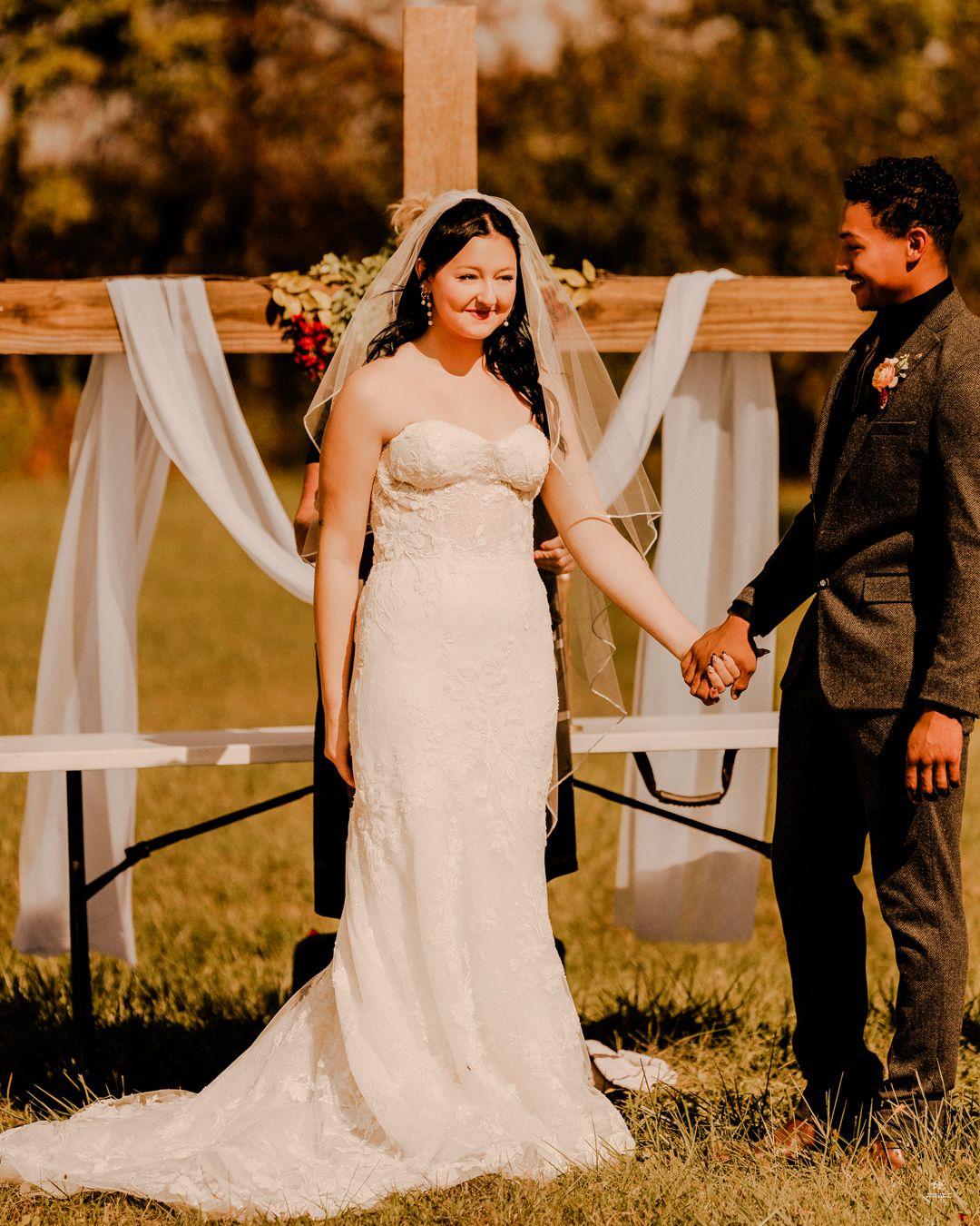 Bride in strapless lace gown holding hands with groom in dark suit before wooden cross altar.