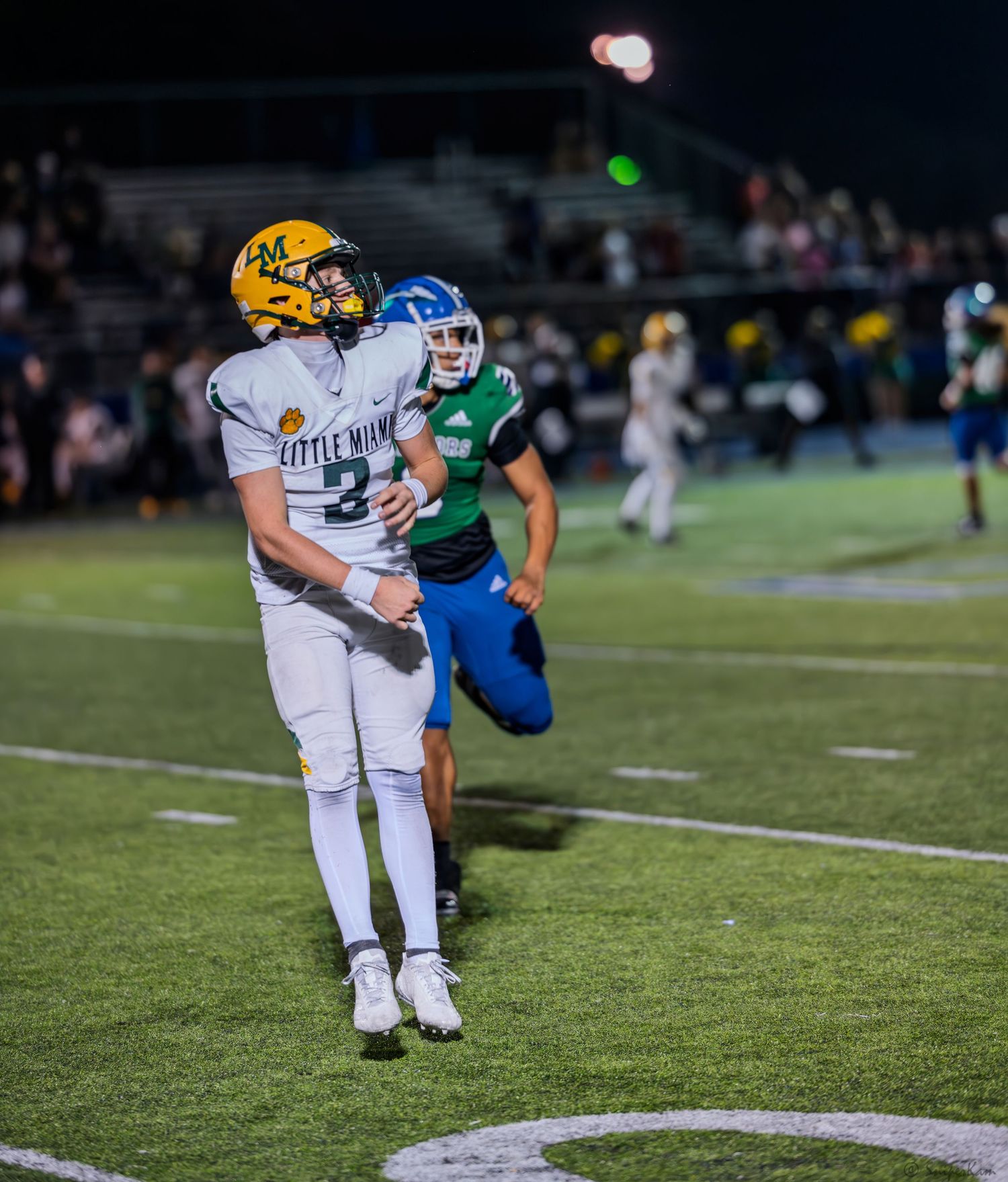 A football player wearing number 3 in white and gold Little Miami uniform throws a pass during a night game.