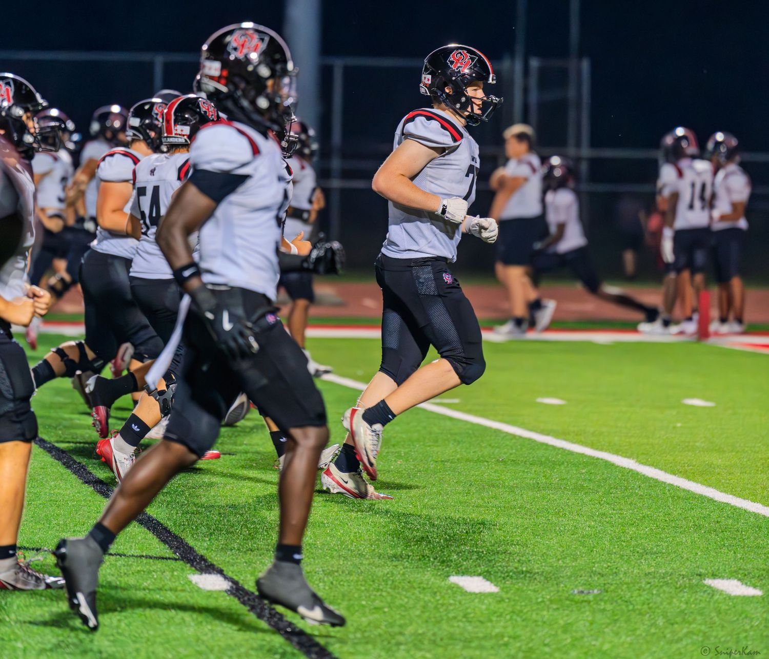 Opposing team players jog along the sideline during a night high school football game on green turf.