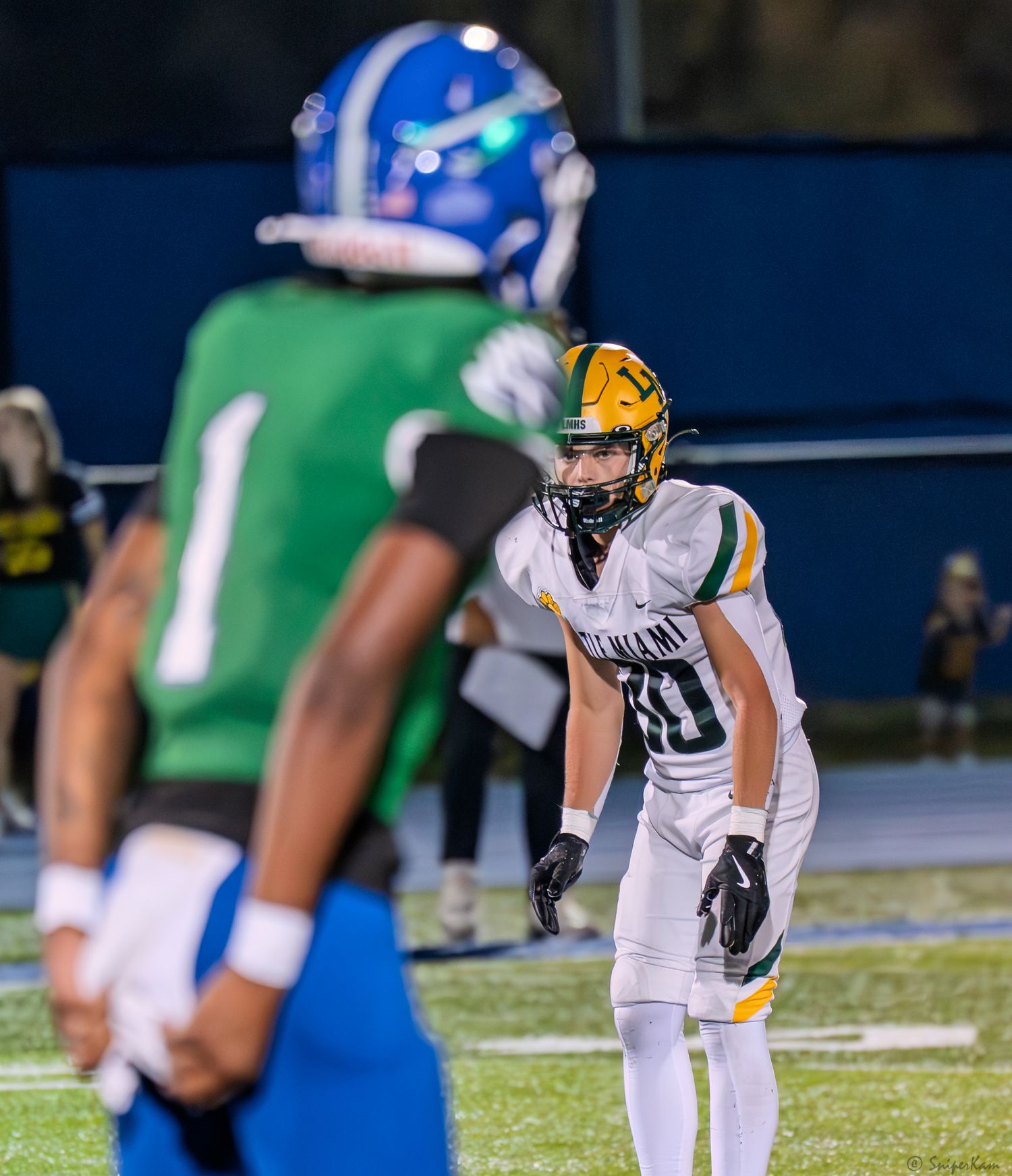 A football player in white and green uniform number 30 lines up against opponent in blue helmet during night game.