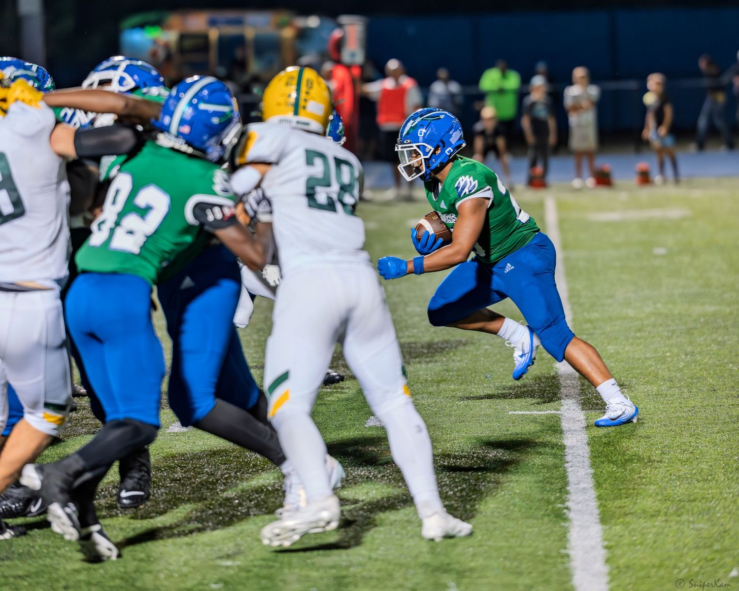 High school football player in green and blue uniform runs the ball during a night game.