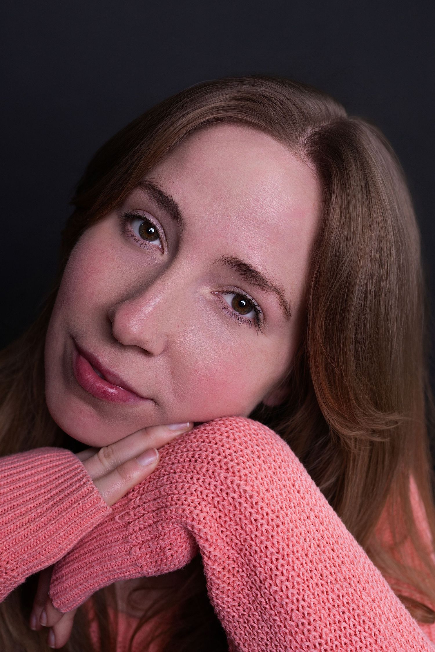Young woman in pink knit sweater resting chin on hands, looking up softly against dark background.