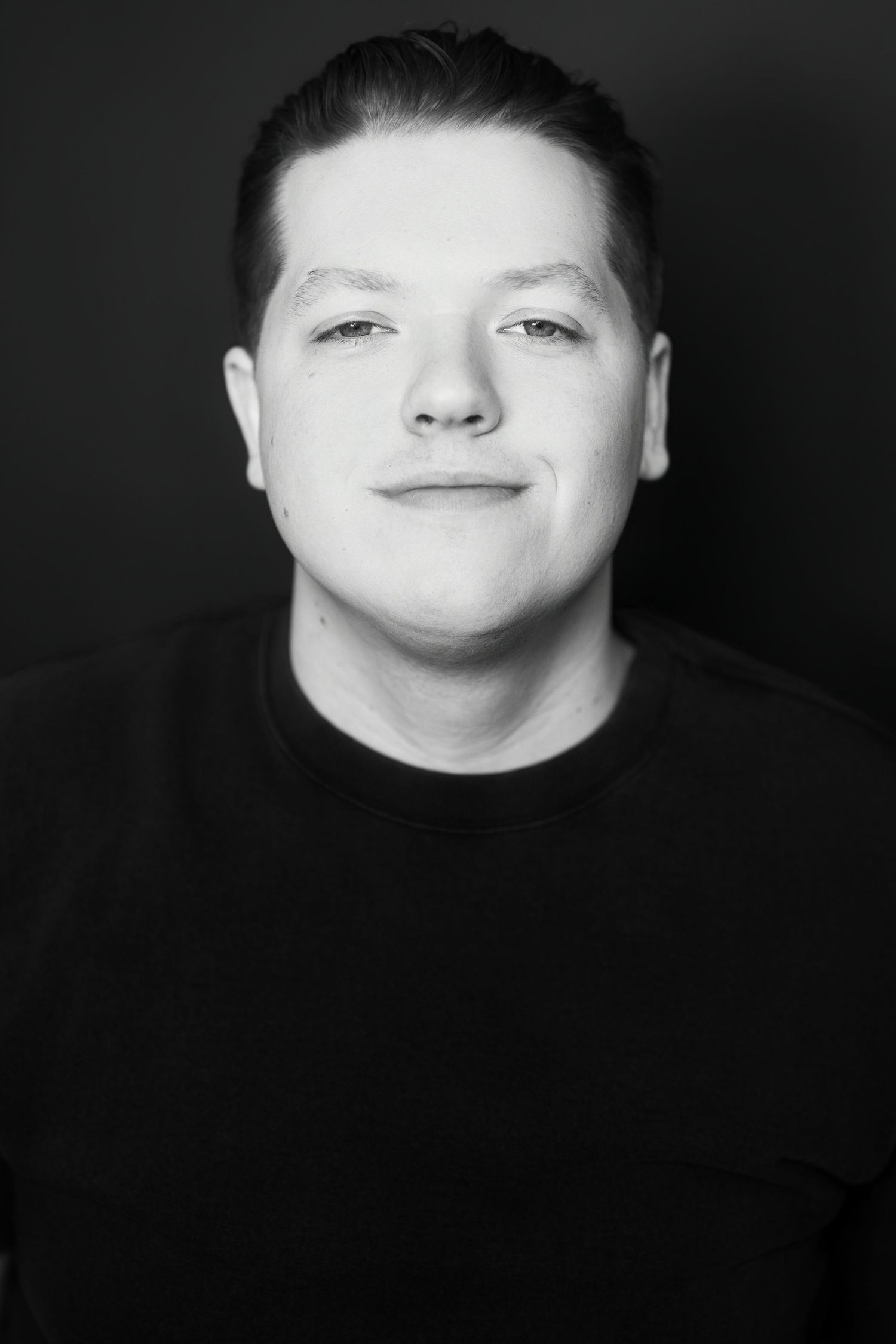 Black and white headshot of smiling young man in black crewneck against dark background.