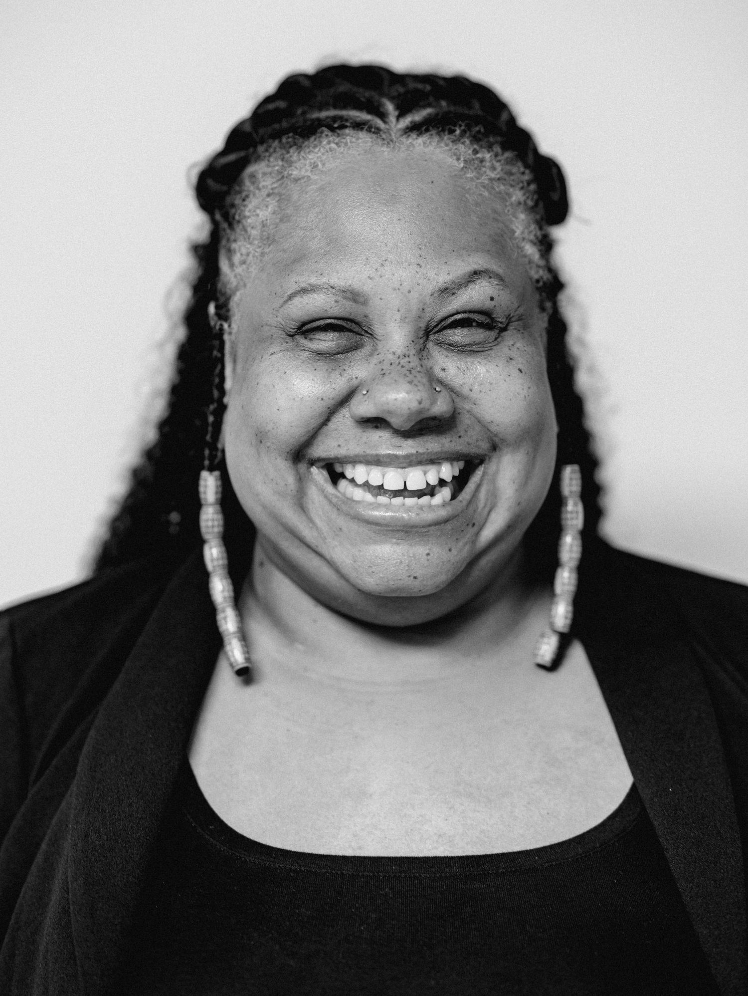 A Black woman with braided hair and dangling earrings smiles widely in a black and white close-up portrait.