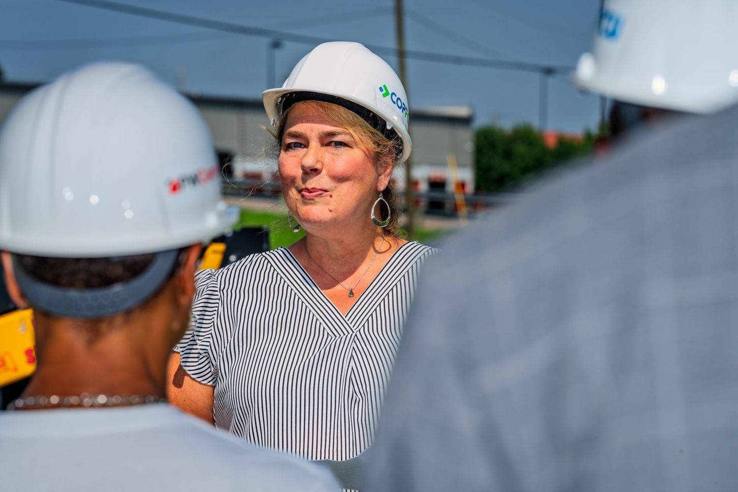 Woman in white COPCU hard hat smiling during outdoor groundbreaking ceremony on a sunny day.