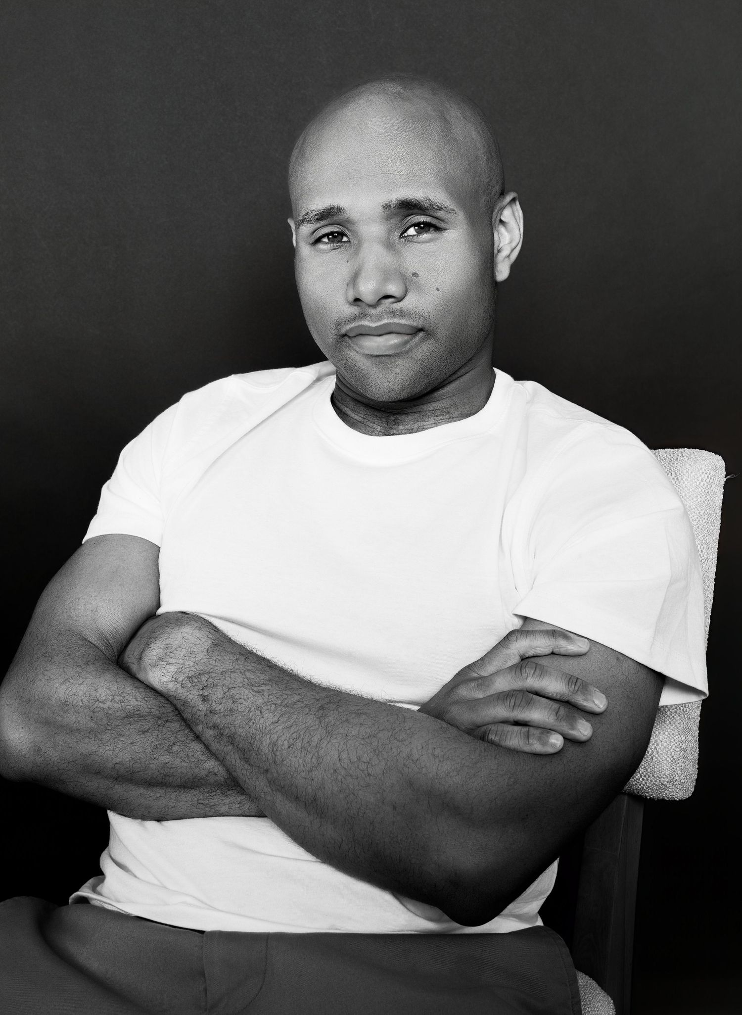 Black and white portrait of bald man in white t-shirt sitting with arms crossed.
