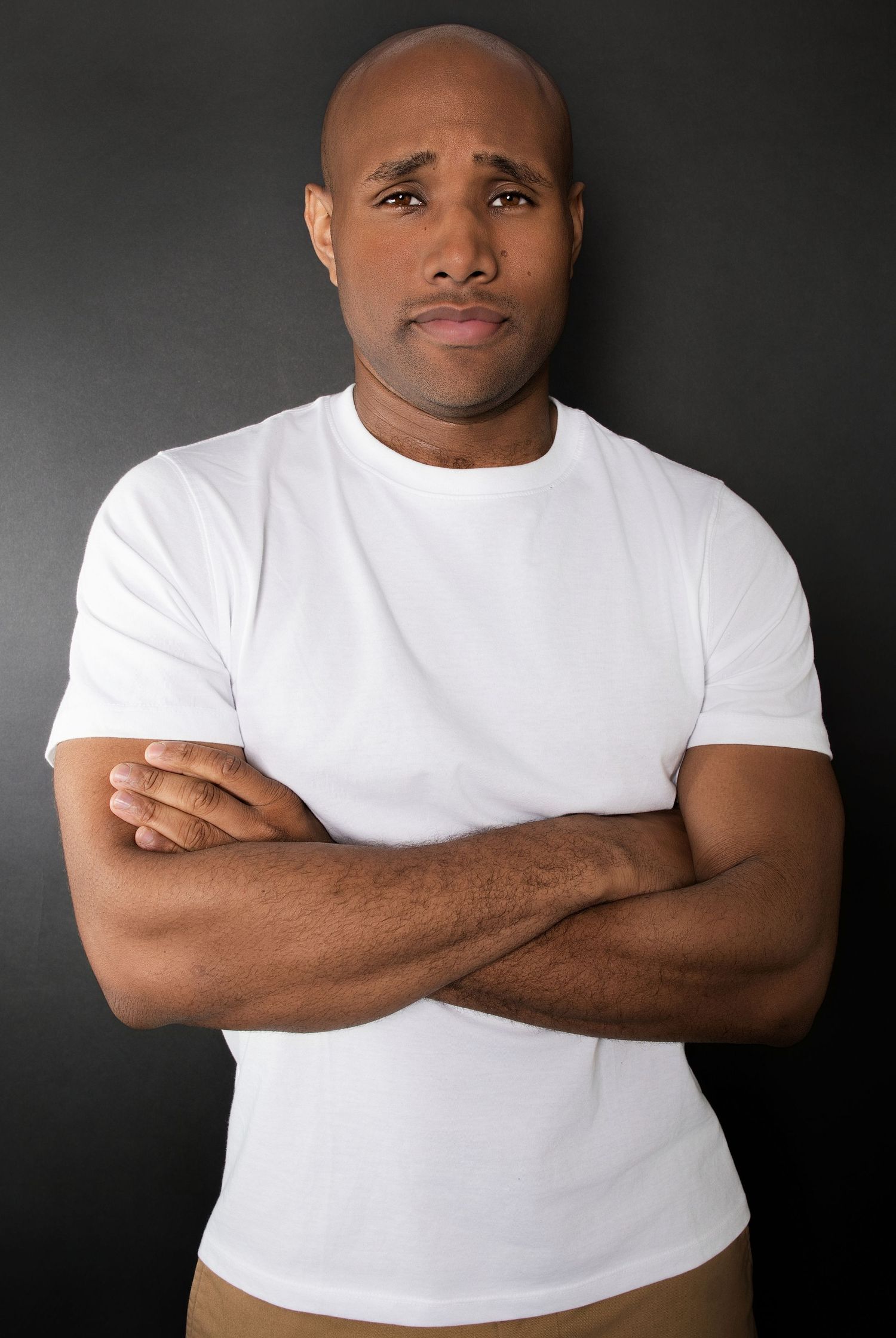 Bald Black man in white t-shirt with worried expression standing with arms crossed in studio.
