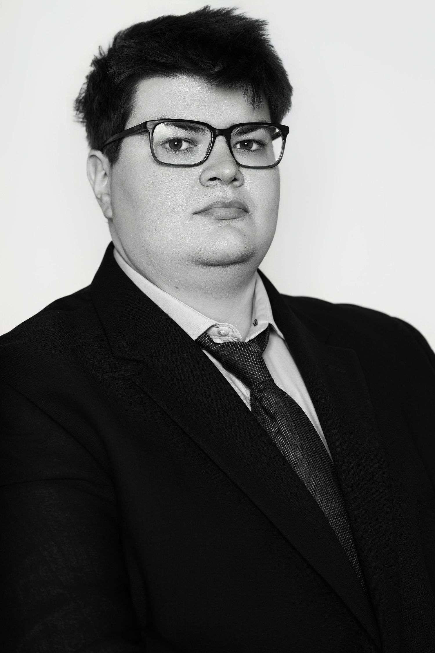 Black and white portrait of a young person in suit, tie, and glasses against white background.