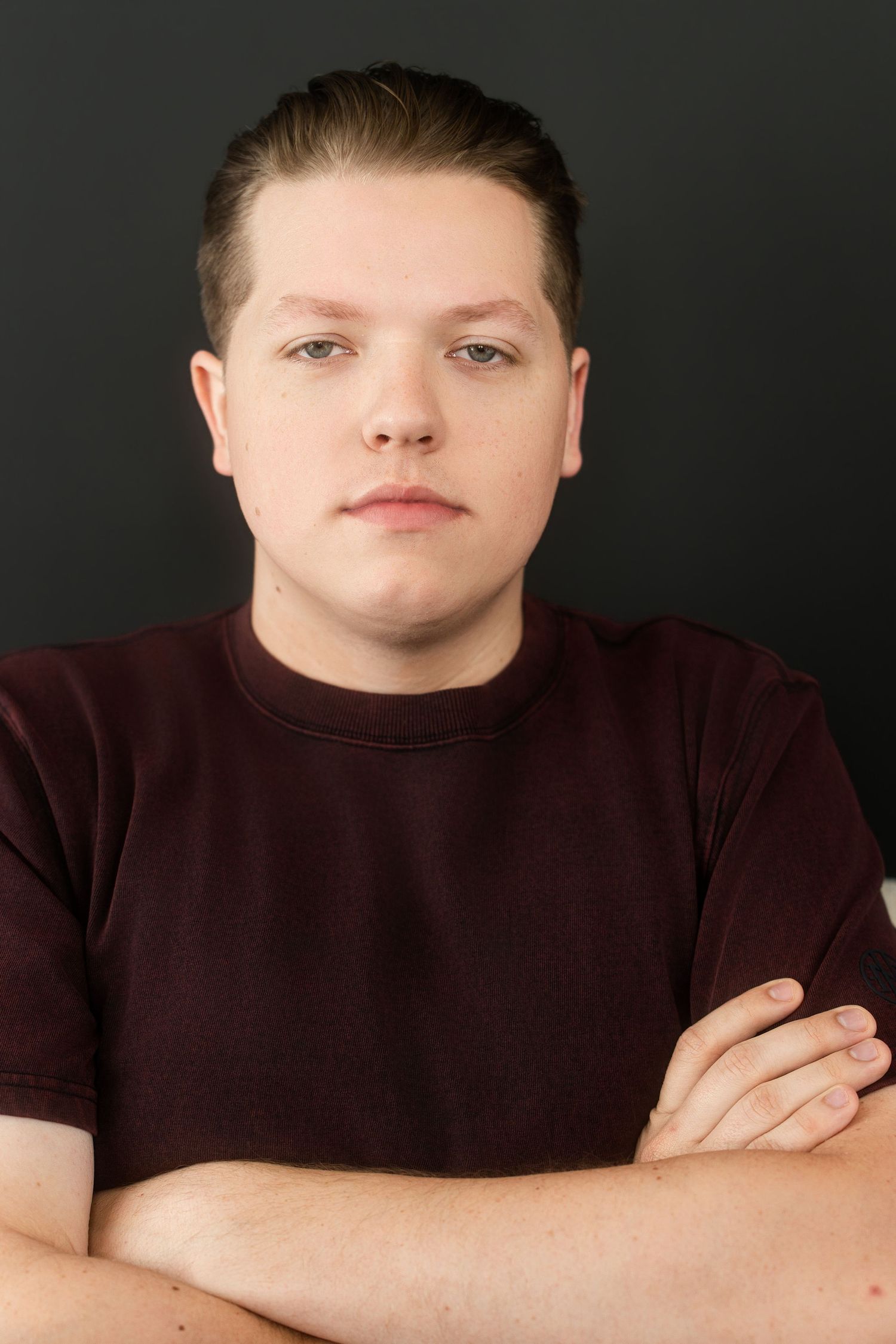 Young man in maroon t-shirt with arms crossed against dark background, serious expression.