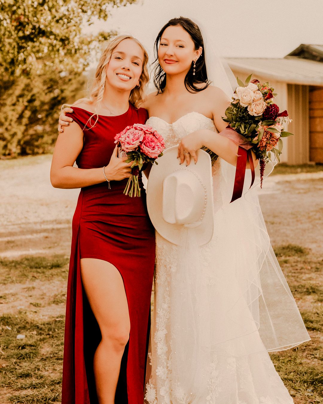 Bridesmaid in red dress and bride in white lace gown holding bouquets at outdoor wedding.