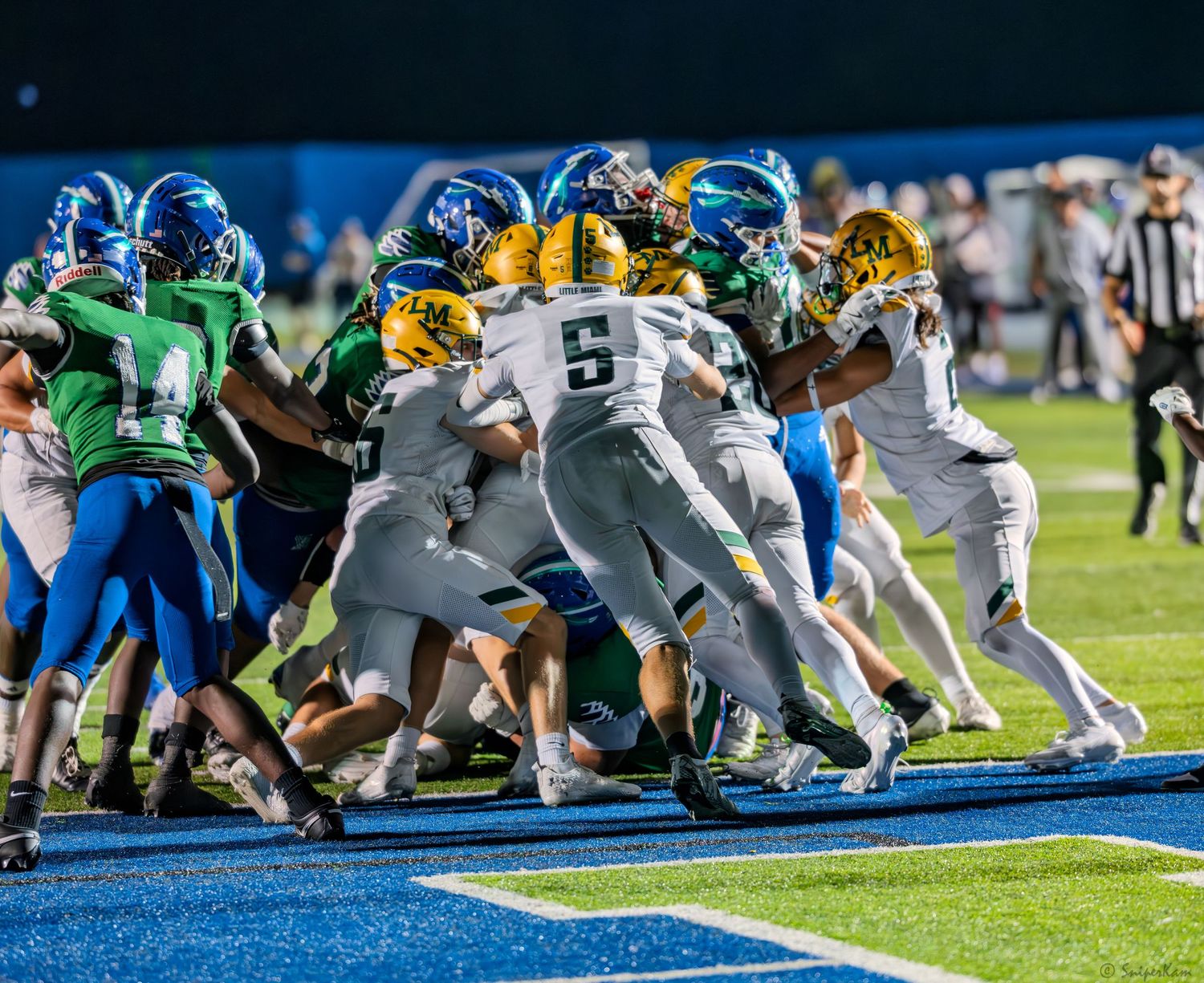 High school football players in blue, green, and gold uniforms collide in an intense goal-line pile-up.