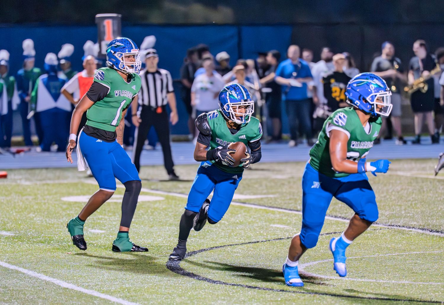 Three Warriors football players in blue and green uniforms run with ball during night game.