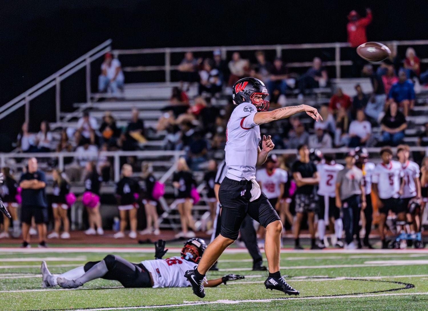 High school quarterback releases pass during night football game with teammates visible on field.