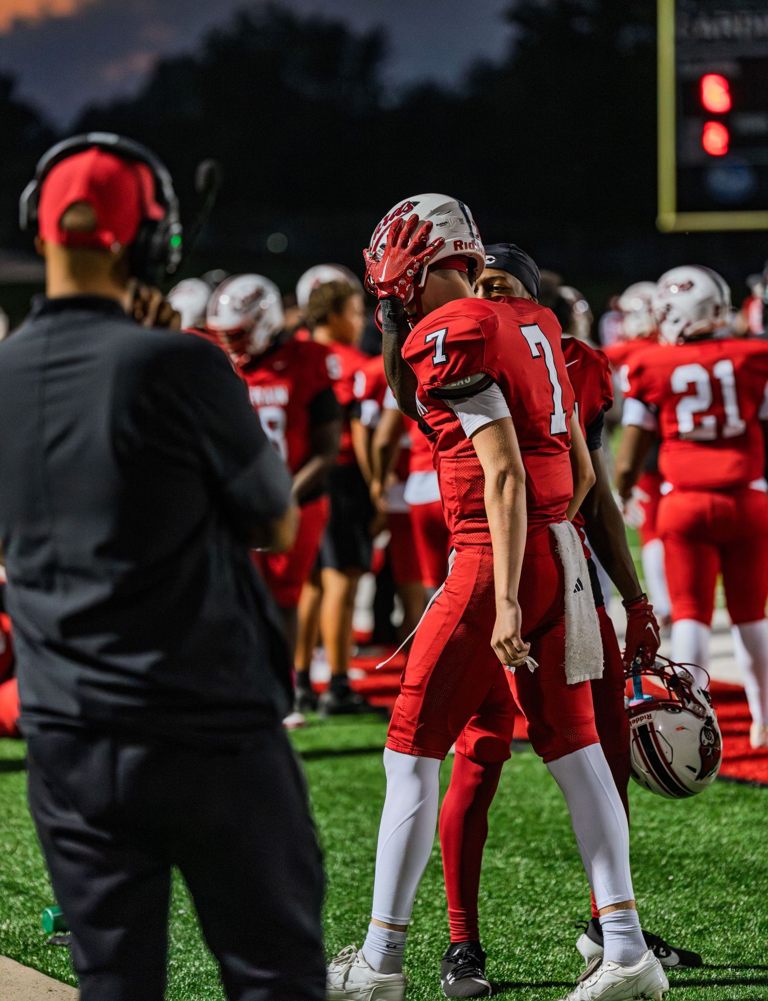 Football player #7 in red uniform walks on sideline holding helmet during a night game.