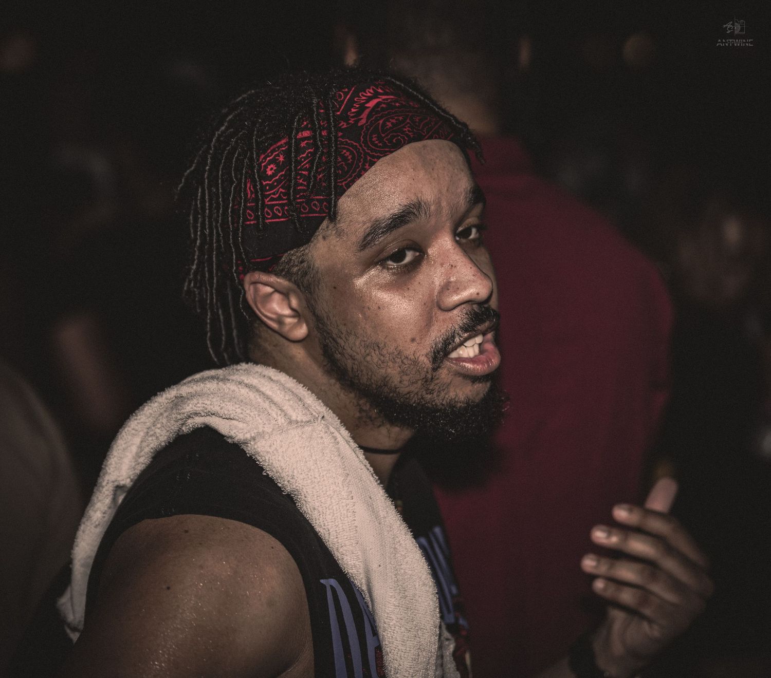A Black man with dreadlocks and a red bandana, towel draped over shoulder, at a lively indoor event.