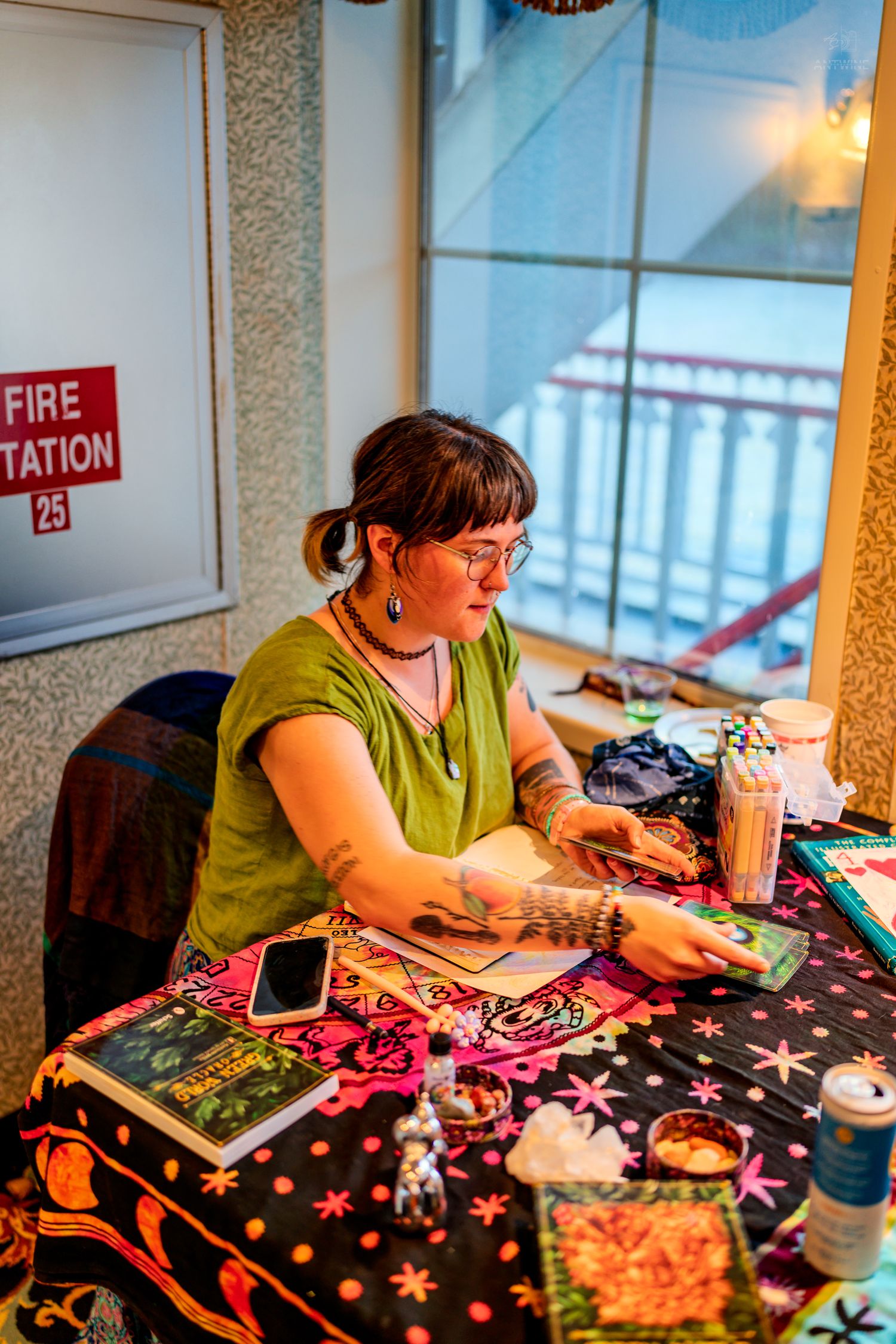 Young woman with glasses reading tarot cards at a colorful table with crystals and books.