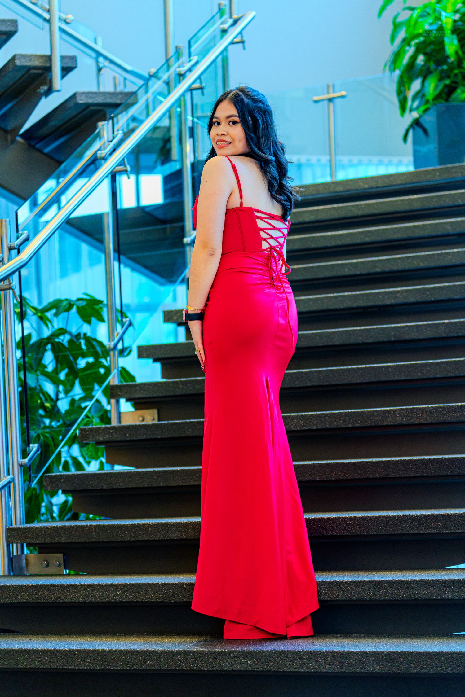 Woman in red mermaid gown with lace-up back posing on stairs at formal venue.