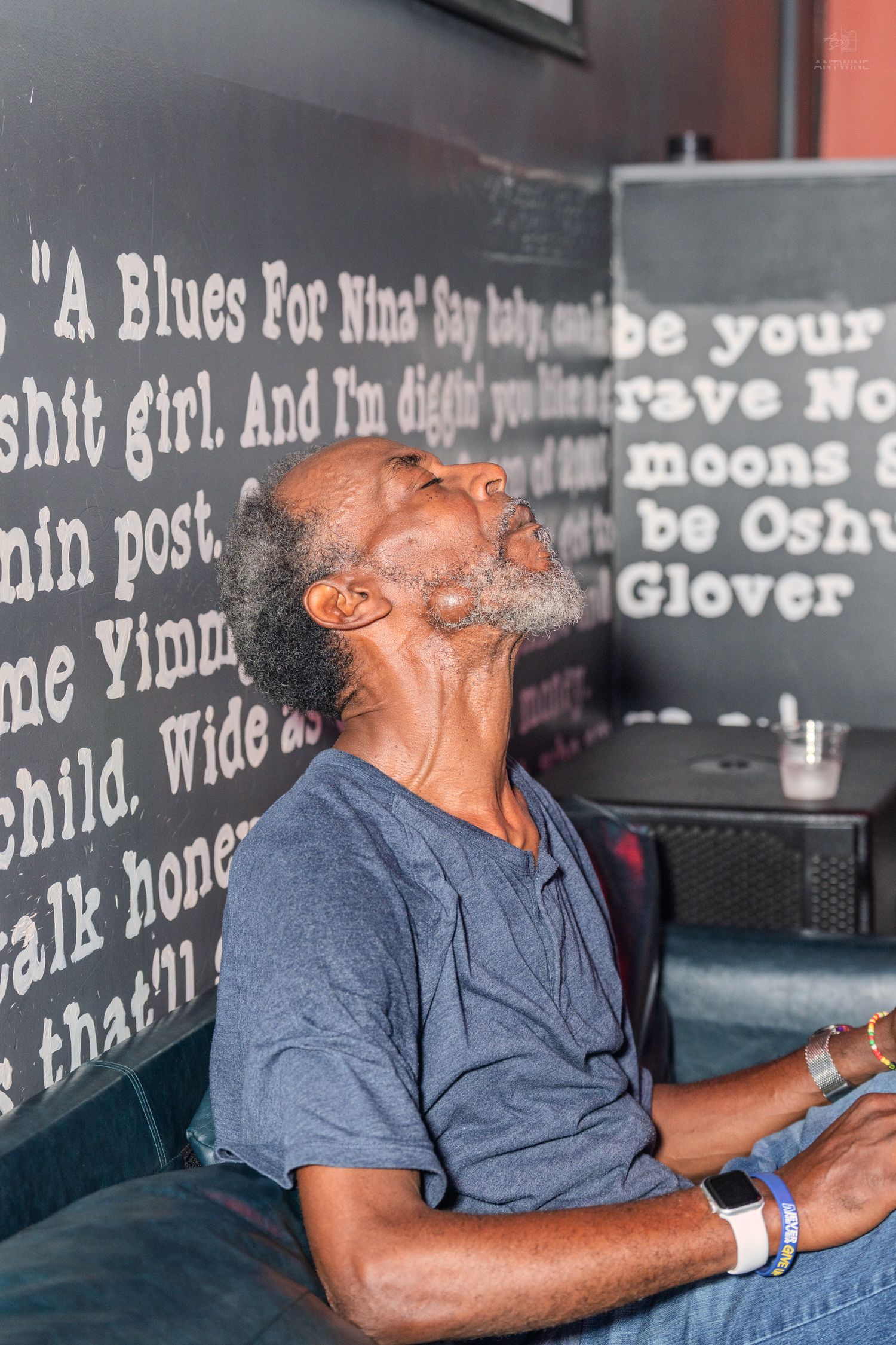 An elderly Black man with a gray beard sits relaxed, head tilted back, in front of a dark wall with blues poetry text.
