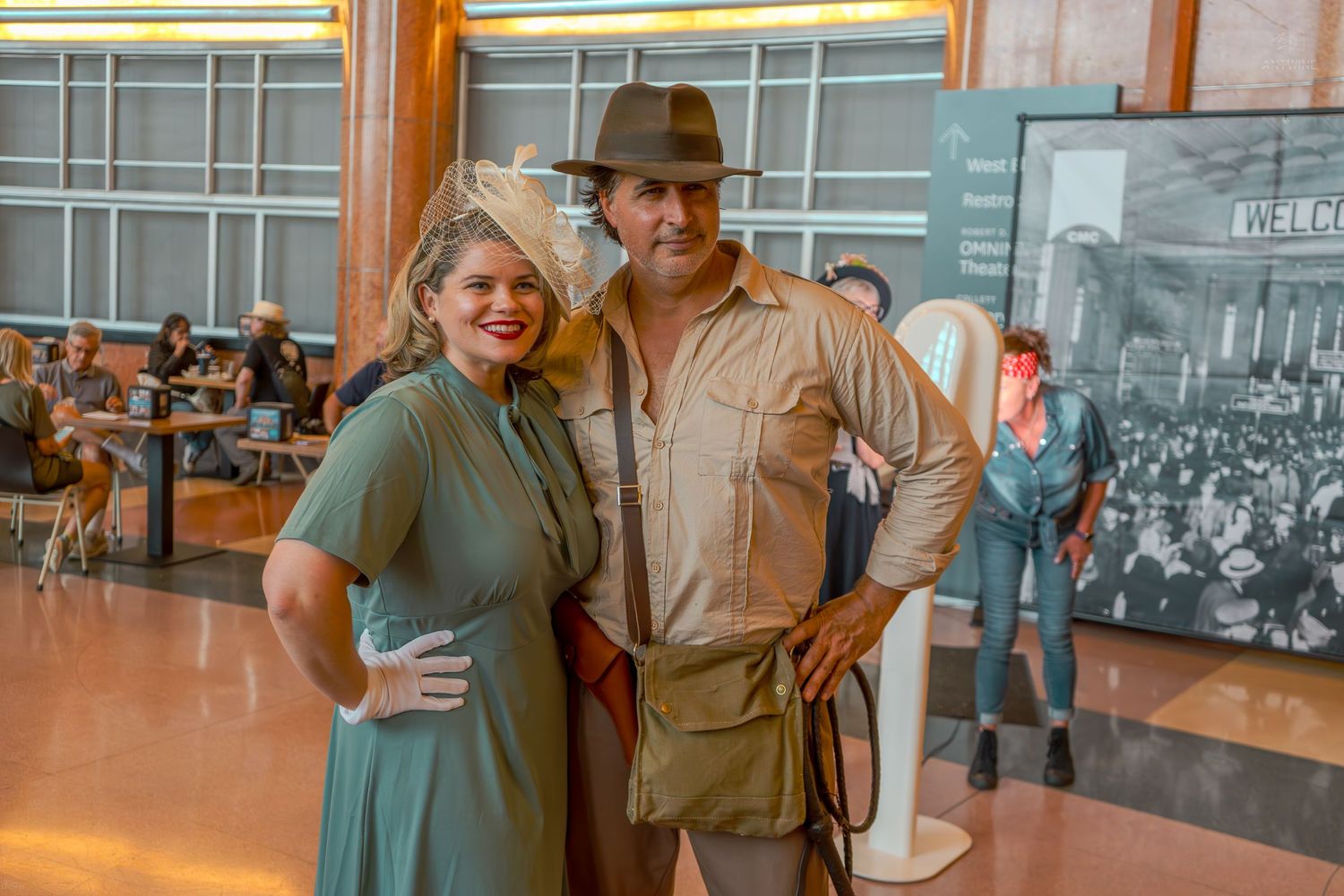 Couple in vintage 1940s costumes, woman in green dress and man as Indiana Jones, posing indoors.