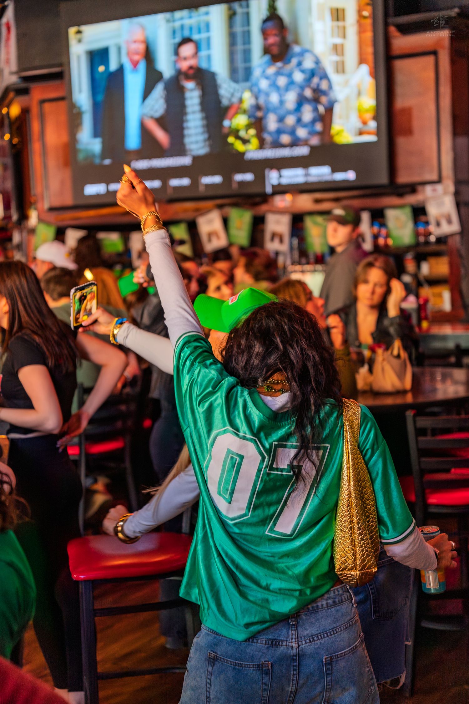 Woman in green #07 jersey raises hand at lively sports bar with TV screen showing movie.