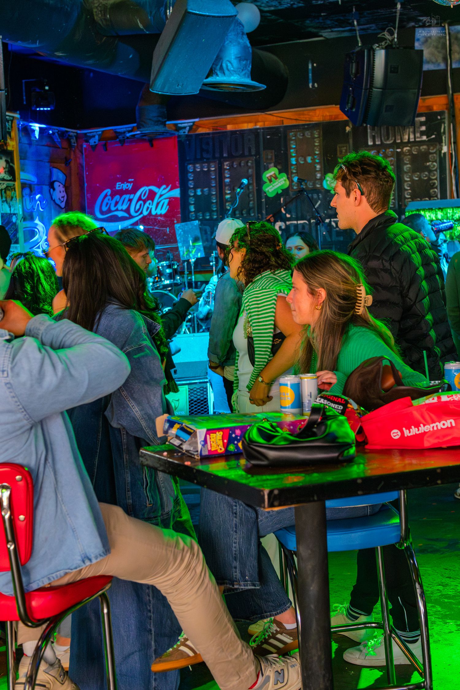 People socializing at a lively bar with Coca-Cola sign, green lighting, and a lululemon bag on the table.