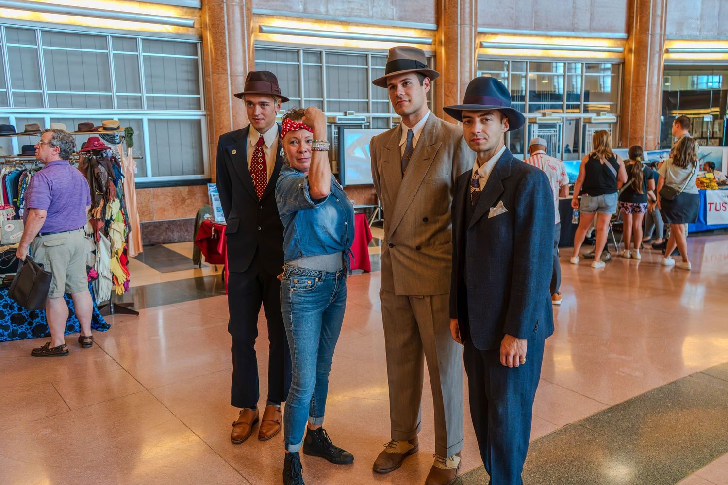 Three men in vintage fedoras and suits pose with a woman in jeans at an indoor retro-themed event.