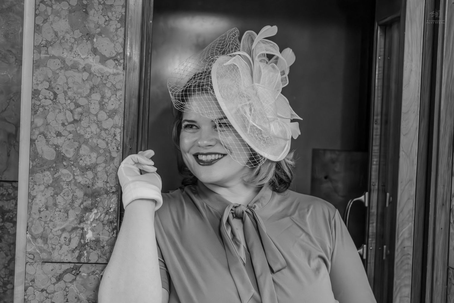 Woman in vintage fascinator hat with floral detail and birdcage veil, smiling in black and white photo.