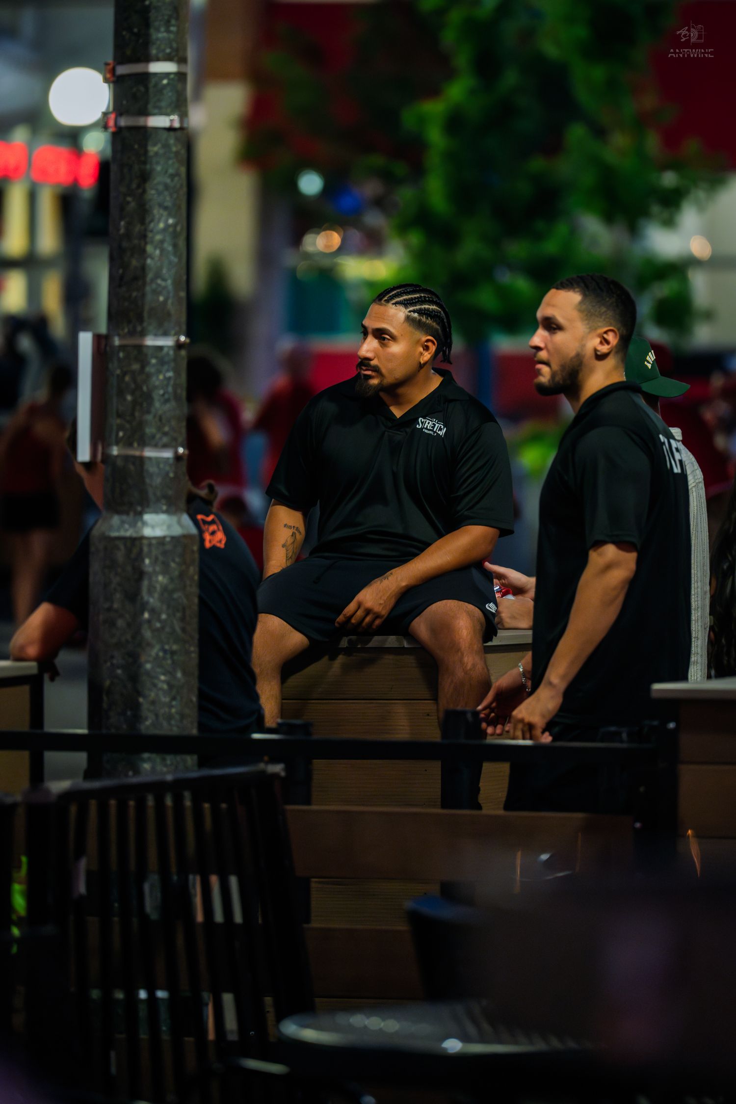 Two men in black shirts sit outdoors at night near a street lamp in an urban setting.