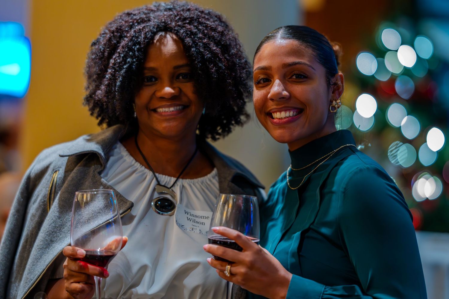 Two smiling women holding wine glasses at a festive holiday party with Christmas tree lights in background.