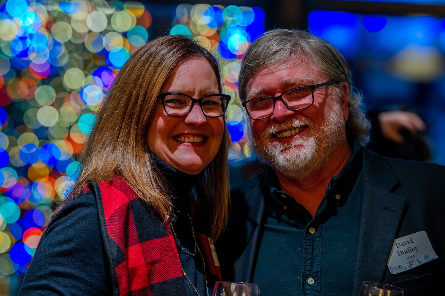 A smiling couple poses together at a festive holiday party with colorful bokeh lights in the background.