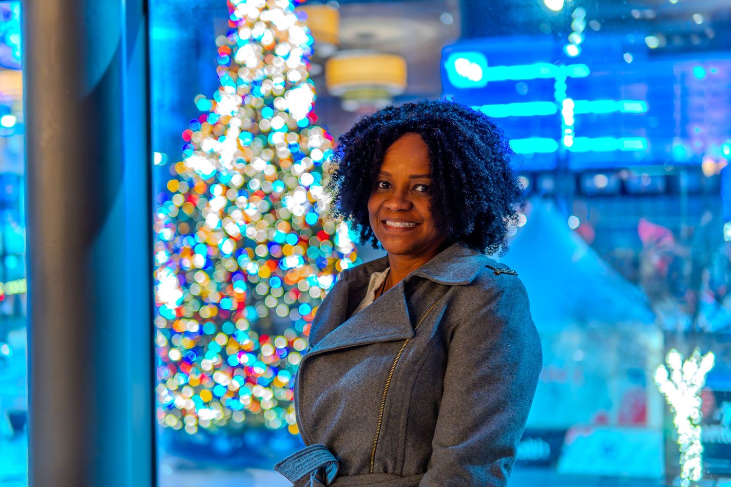 Woman smiling near a decorated Christmas tree with colorful bokeh lights in a festive holiday setting.