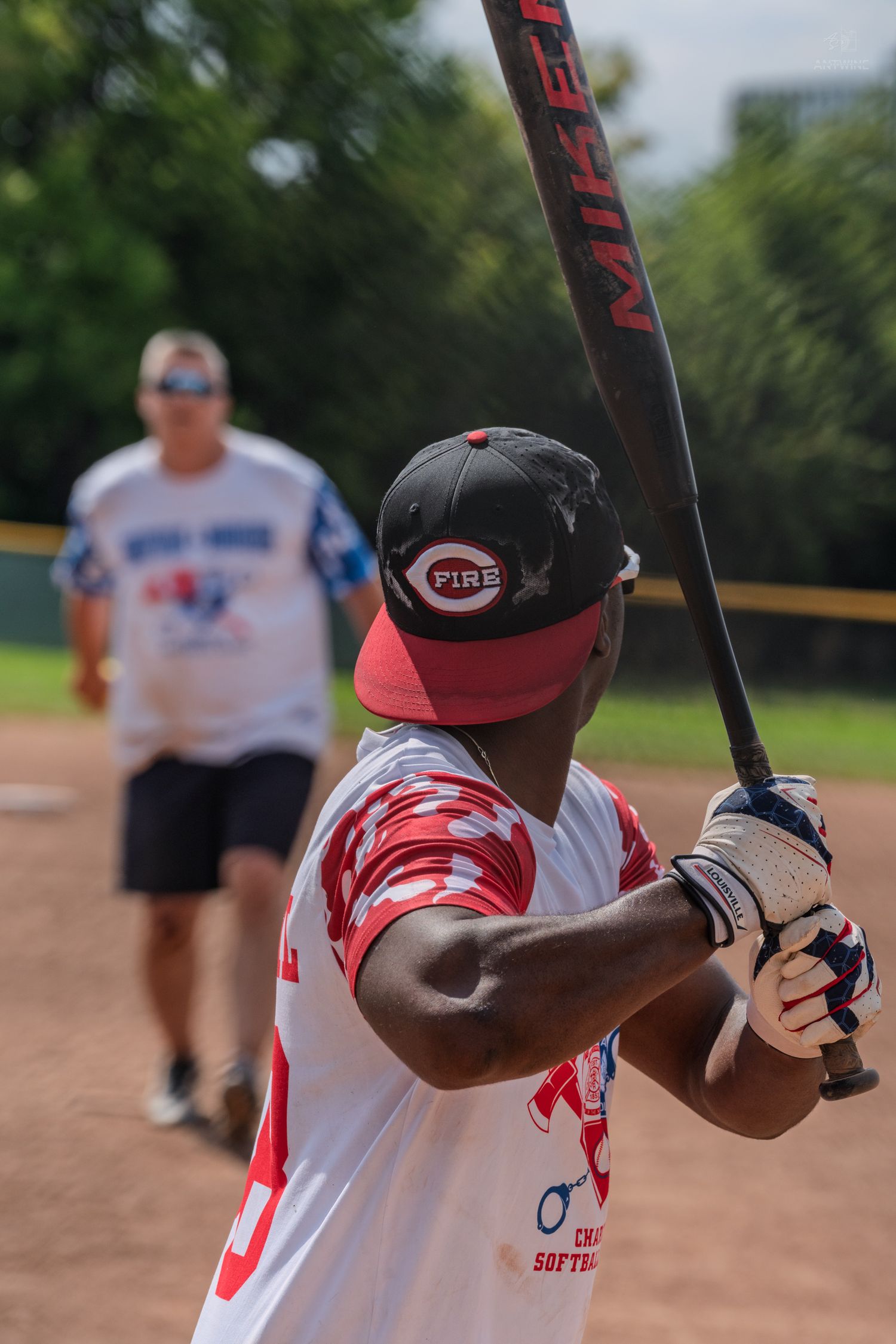 Batter holding a Miken bat at a charity softball game wearing a Fire team cap and red camo jersey.