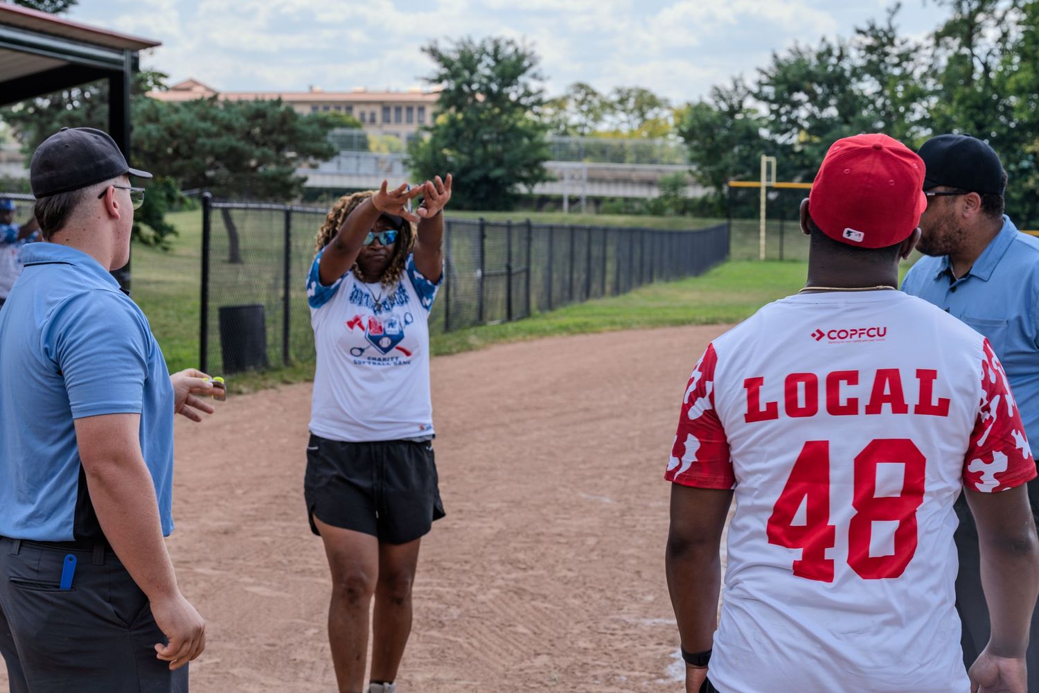 A woman gestures while talking to players in baseball uniforms on a dirt field during an outdoor sports event.