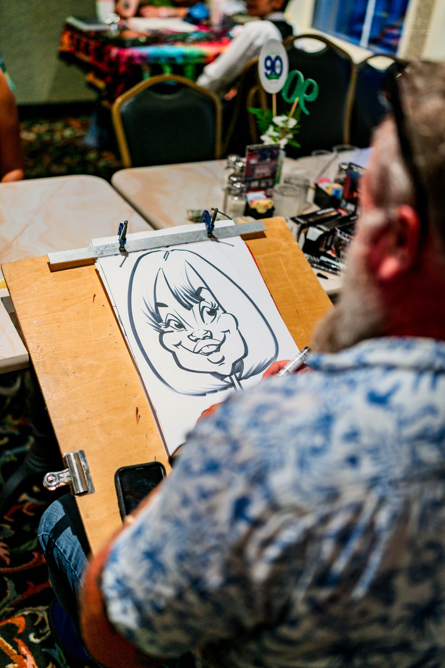 Artist draws an exaggerated caricature portrait on a drawing board at an indoor event.