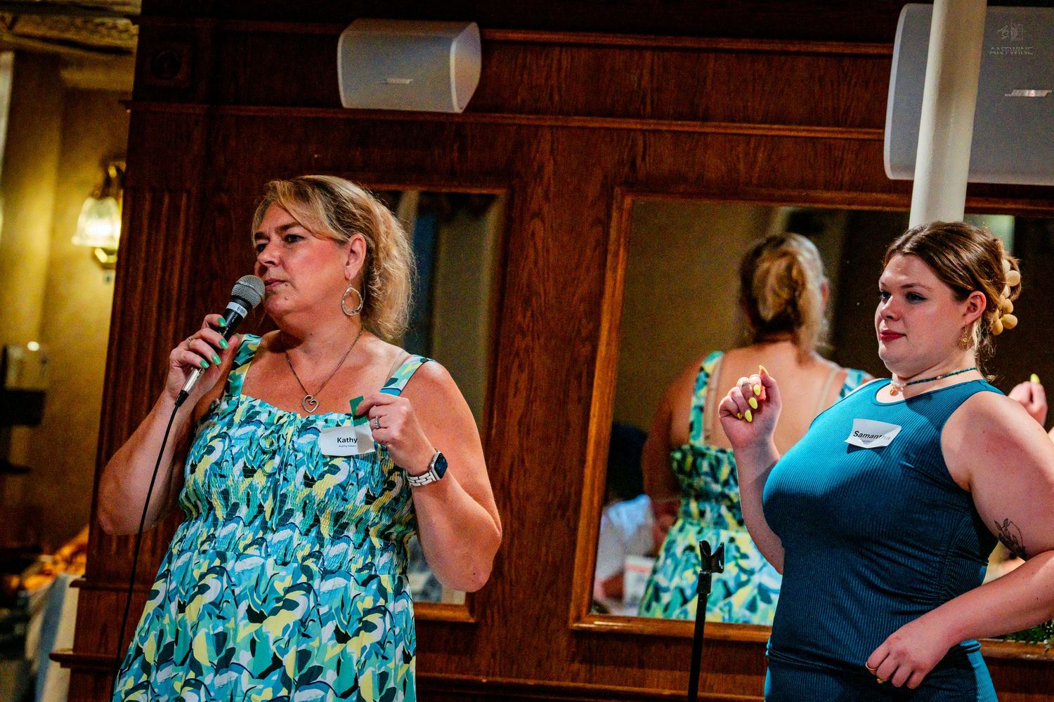 Two women, one speaking into a microphone wearing a floral dress, present at an indoor banquet event.