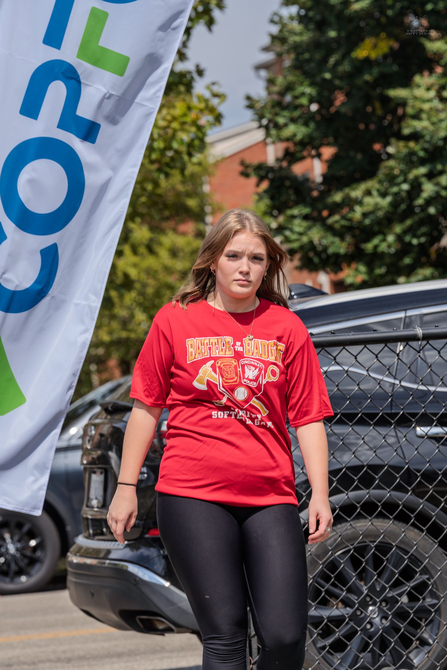 Young woman in red Battle of the Badges softball shirt walking near a police vehicle.