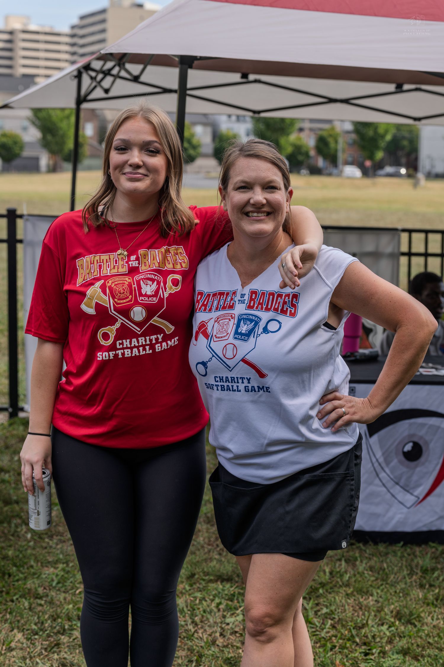 Two women smile at a charity softball game, wearing red and white event t-shirts outdoors.