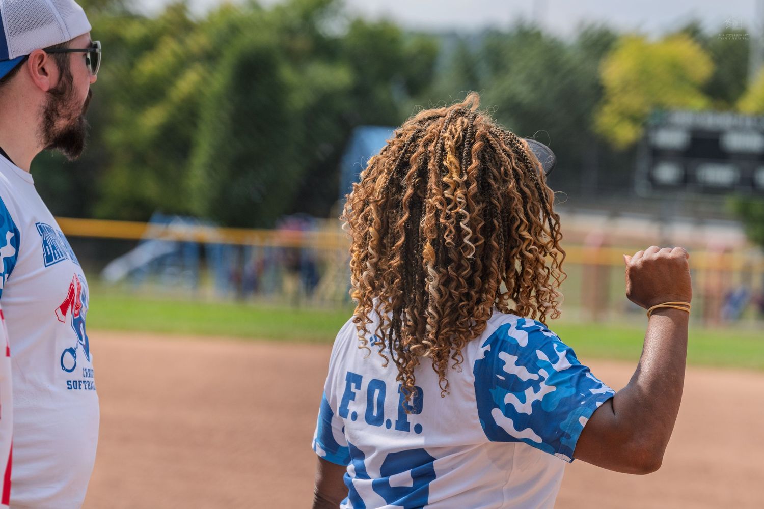 Woman in FOP blue jersey raises fist in celebration on softball field sideline.